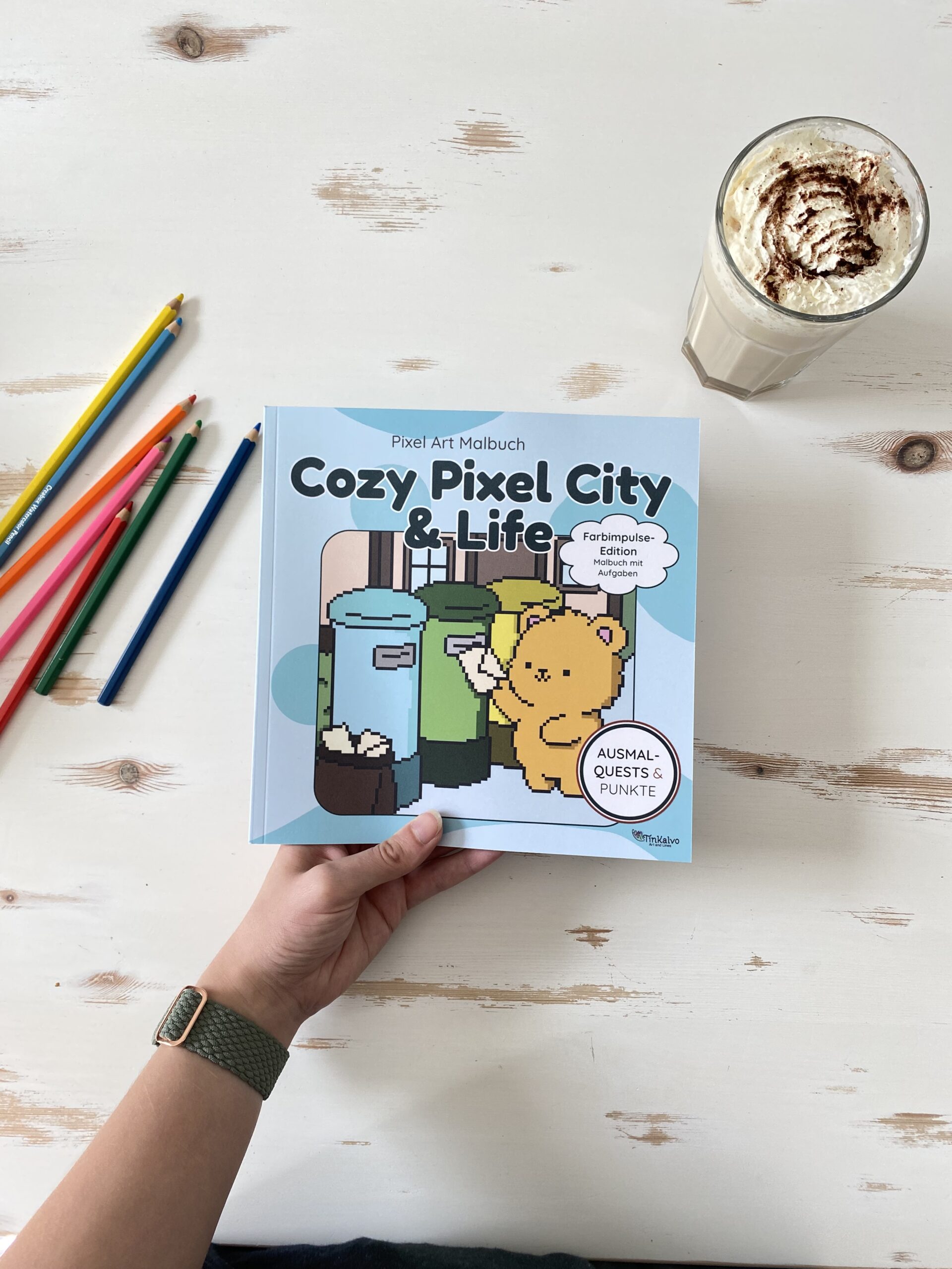 Malbuch Cozy Pixel City Farbimpulse Edition in Hand gehalten mit Stiften und Getränk im Hintergrund